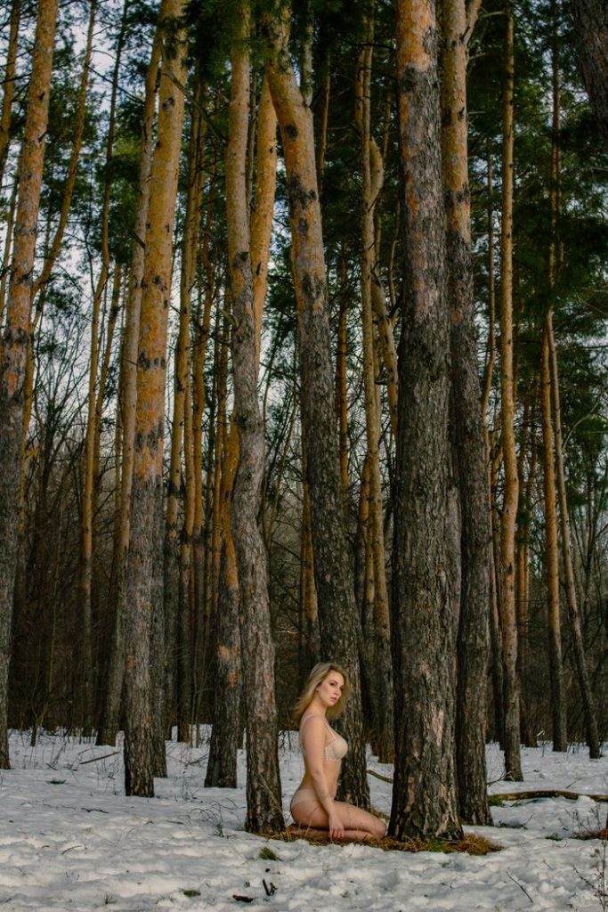 A woman kneeling in a snowy forest among tall pine trees, capturing nature's serenity.