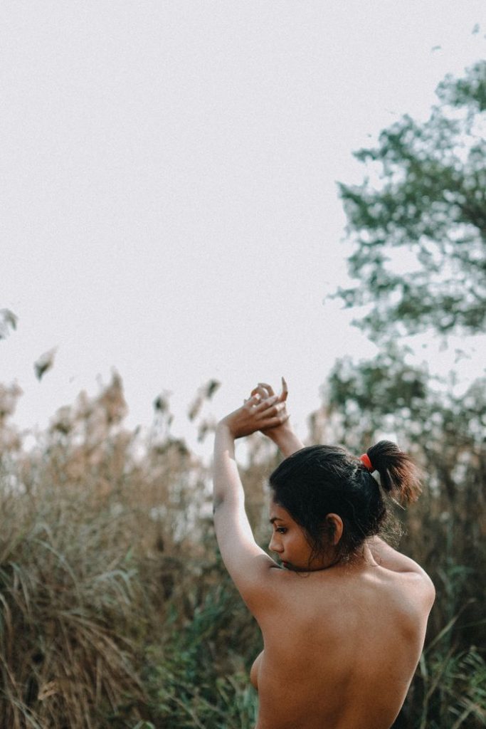 Back view of faceless topless female standing near field with dry grass and trees with raised arms in daylight in nature